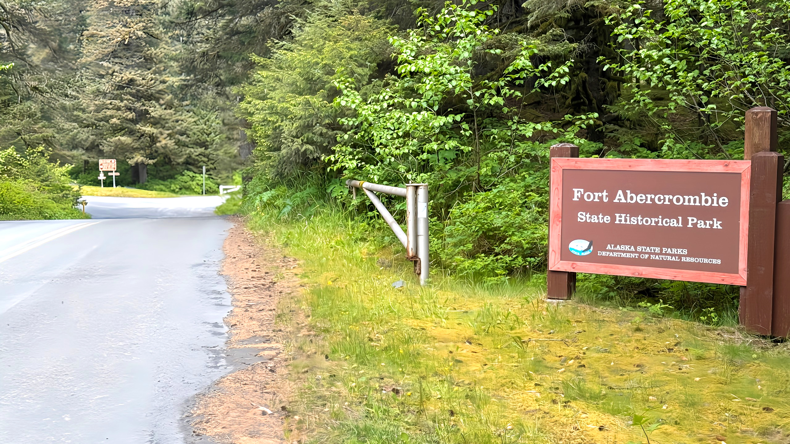 A wooden sign reading Fort Abercrombie State Historical Park stands by a forested Kodiak roadside, with green foliage and a nearby trail in the background.