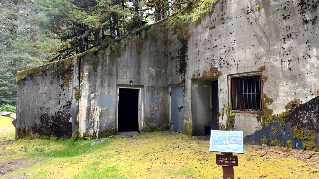 A moss-covered concrete bunker with two open doorways and one barred window sits amid Kodiak’s lush trees and greenery. A small informational sign stands in front of the structure.