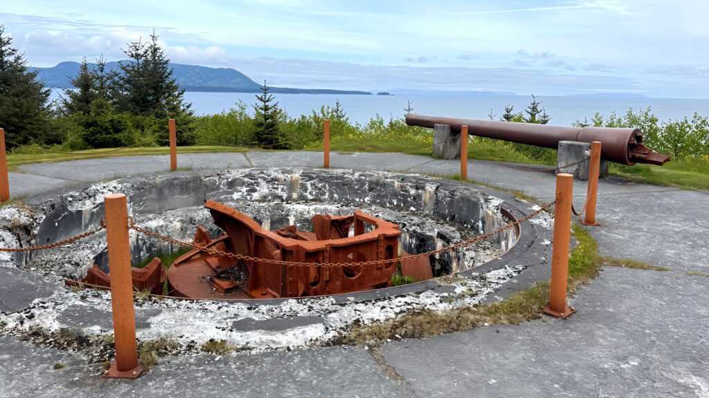 A rusted artillery gun and circular concrete gun emplacement overlook the Kodiak sea, with trees and mountains in the background under a cloudy sky. Orange posts and chains surround the weathered structure.