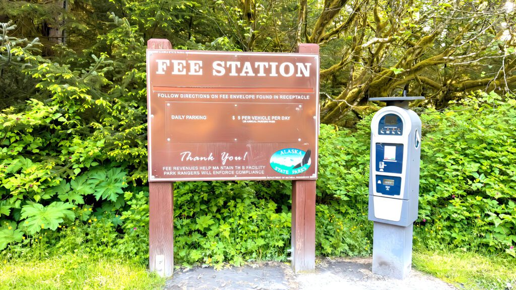 A brown Fee Station sign with instructions, daily parking fees, and an Alaska State Parks logo stands next to a parking payment machine surrounded by green foliage and trees on Kodiak Island.