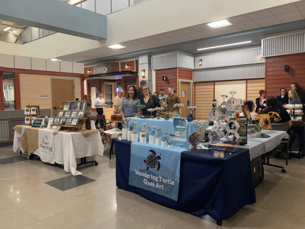 Indoor craft fair featuring local art and crafts Kodiak, with vendor tables displaying handmade items like glass art and framed pictures. Two women smile behind a table labeled Wandering Turtle Glass Art as shoppers browse in the background.