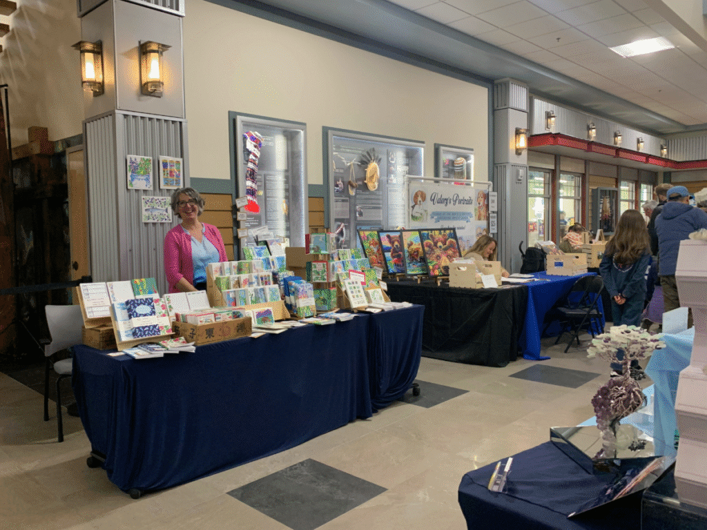 A woman stands behind a table filled with colorful local art and crafts from Kodiak at an indoor market or craft fair. Other vendors and displays line the hallway as shoppers browse nearby.