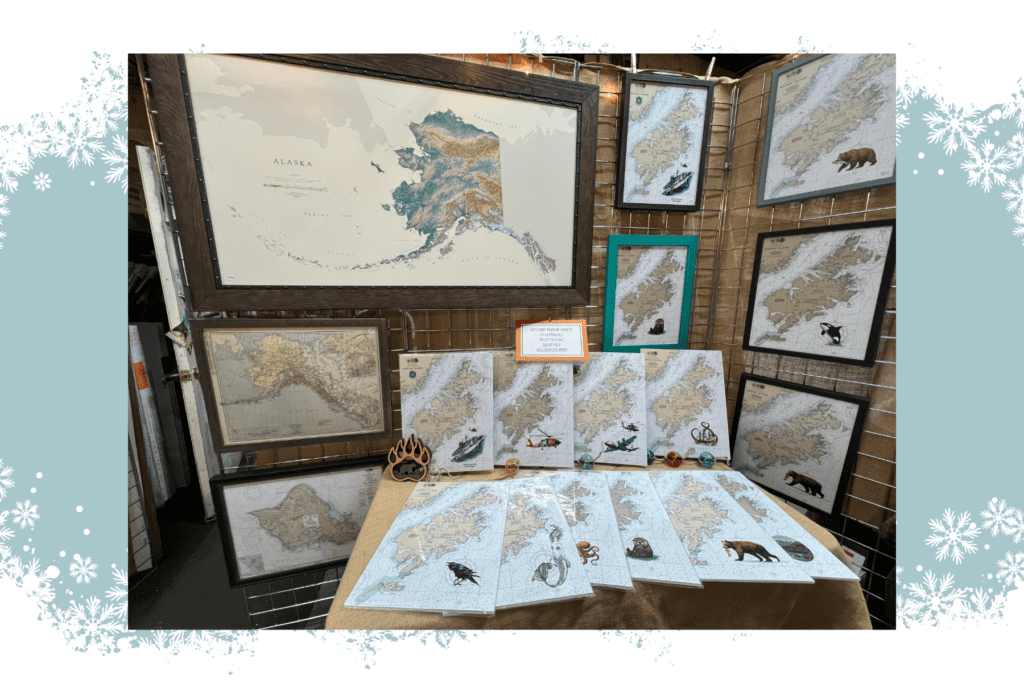 A display of detailed Alaska maps at The Frame Shop, perfect for holiday gifts in Kodiak, features regional artwork and wildlife illustrations. A large framed map sits above smaller ones, all surrounded by a snowflake border.