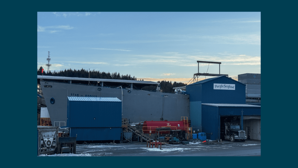 A large gray ship, the Star of Kodiak, is docked beside blue industrial buildings labeled “Pacific Seafood,” with trucks and equipment nearby on a snowy ground under a partly cloudy sky.