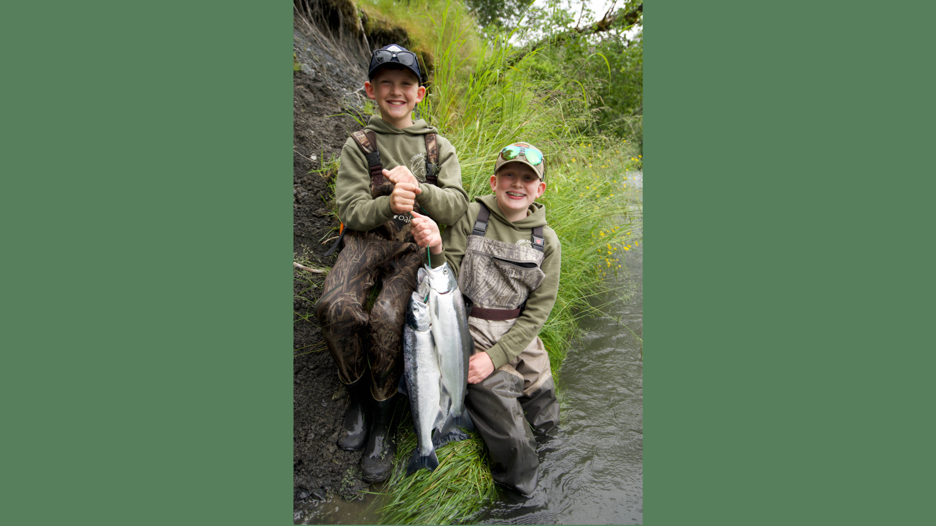 Two smiling children in fishing gear kneel by a stream, proudly holding up a large silver fish they caught. They are surrounded by green grass and water, with one child partially standing in the stream.