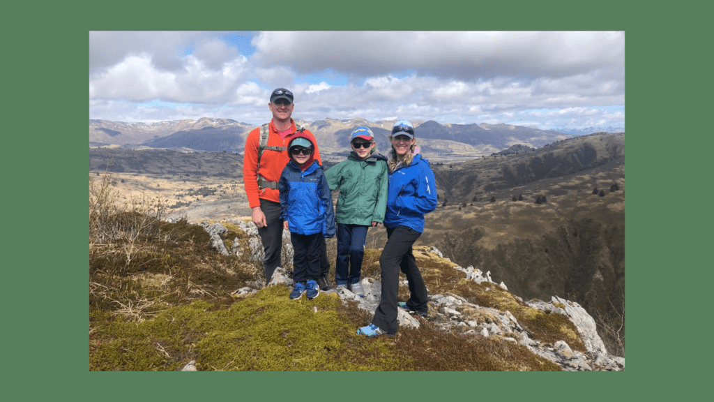 Four people in colorful jackets and hats stand on a rocky hilltop, smiling and posing with scenic mountains behind them. Experience the best of Kodiak with Kodiak Alaskan Adventures as you explore breathtaking views and unforgettable moments outdoors.