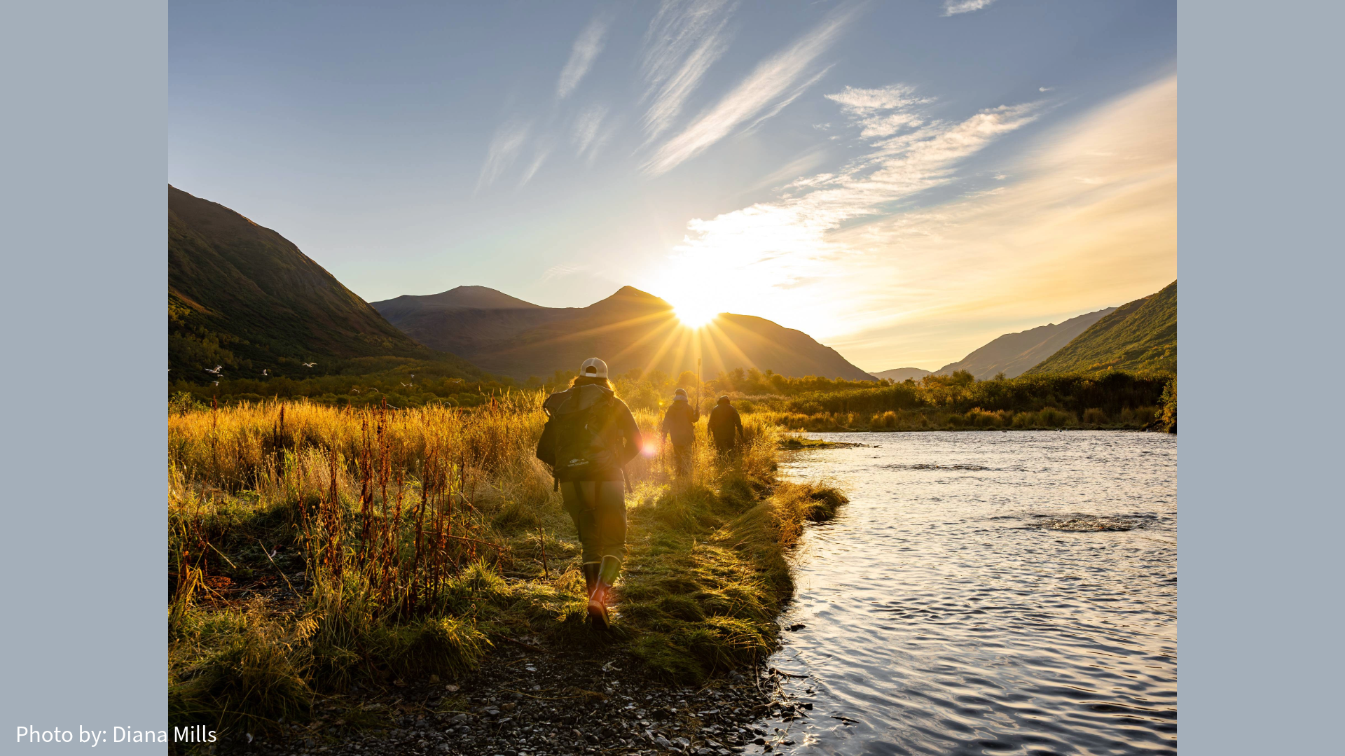 Two people walk along a grassy riverbank toward the mountains at sunrise, golden sunlight illuminating the landscape. A clear river runs beside them under a partly cloudy sky near Kodiak Island Lodges.