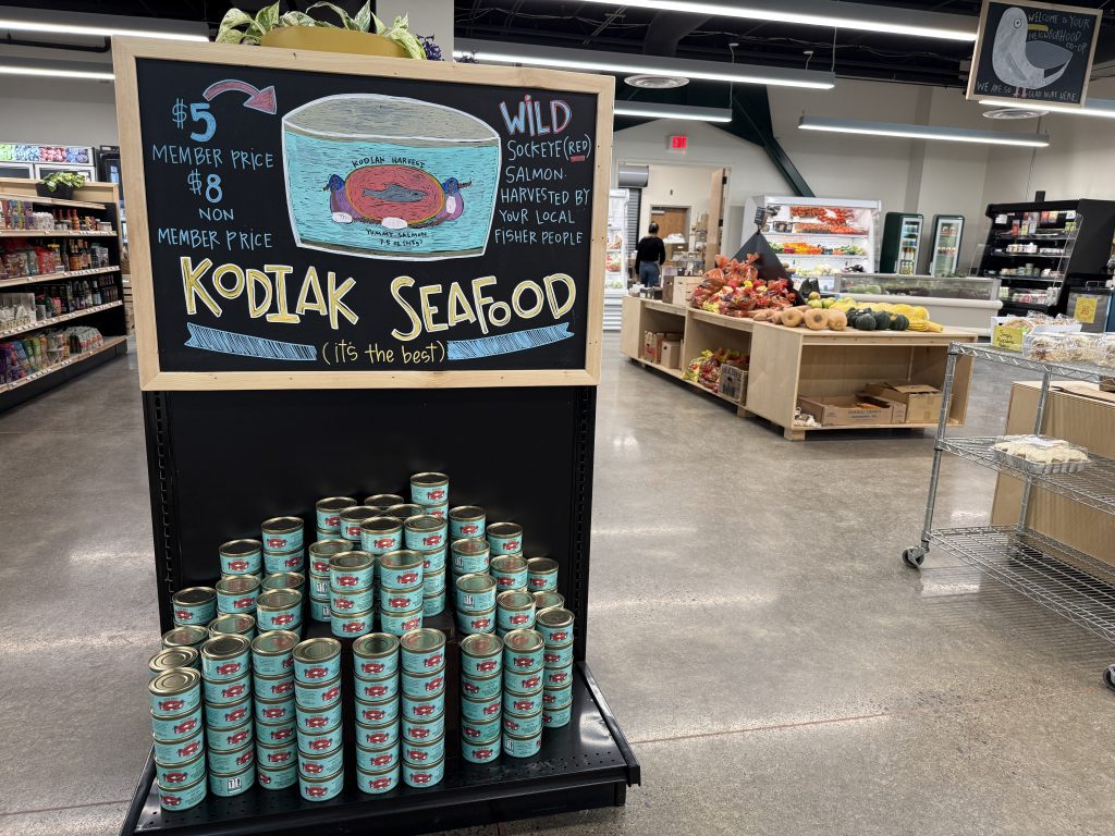 A grocery store display features stacked cans of Kodiak Seafood and a chalkboard sign promoting wild sockeye salmon, with special prices for Kodiak Harvest Food Co-op members. Produce and other grocery items are visible in the background.