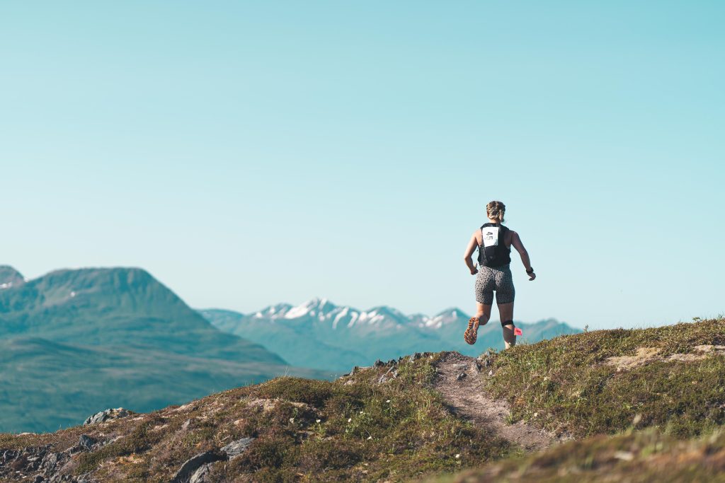 A person runs on a narrow trail over a grassy hill with mountains and snow-capped peaks visible in the background under a clear blue sky.