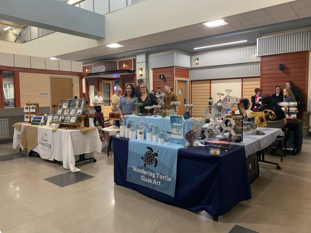 Indoor craft fair at BearTown Market Kodiak with vendors behind tables displaying art, jewelry, and crafts. Two women smile behind a “Wandering Turtle Glass Art” table. Other shoppers and displays are visible in the background.