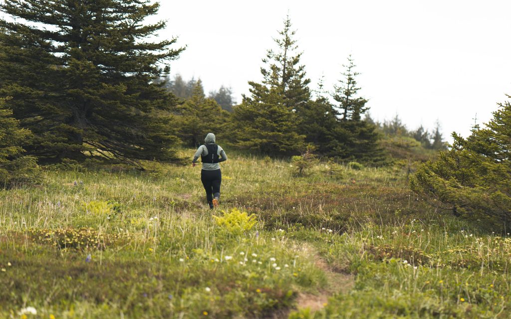 A person wearing a hoodie and backpack runs along a narrow trail through a grassy, flower-dotted meadow surrounded by evergreen trees on a cloudy day.