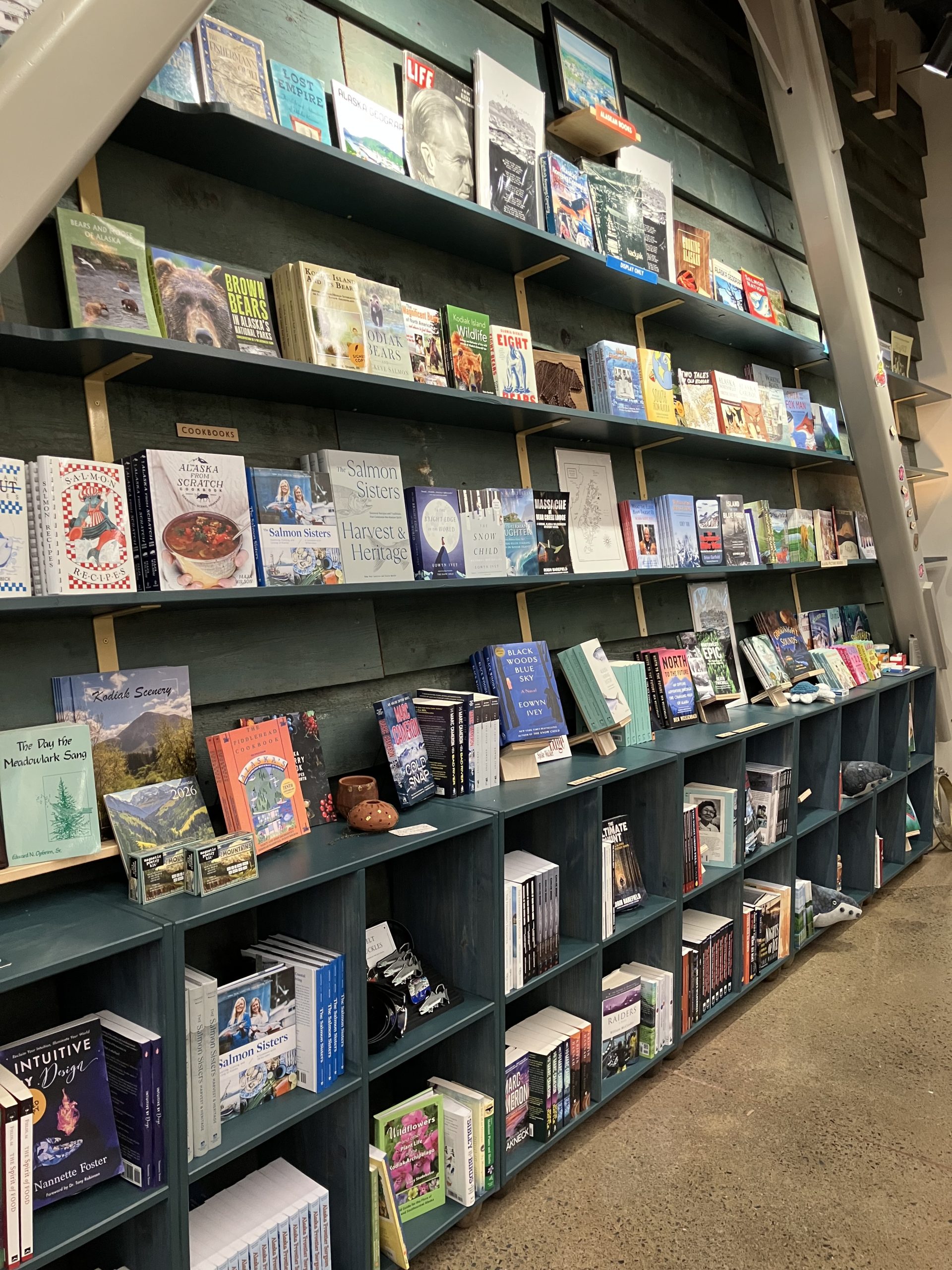 Books are displayed on blue wooden shelves along a green wall in a bookstore, with titles arranged both upright and face-out—creating the perfect cozy spot for browsing during Kodiak rainy days, surrounded by small decorative items and trinkets.
