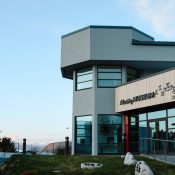 A modern gray building labeled Alutiiq Museum with large windows stands next to a white church with blue domes; mountains are visible in the background under a clear sky.