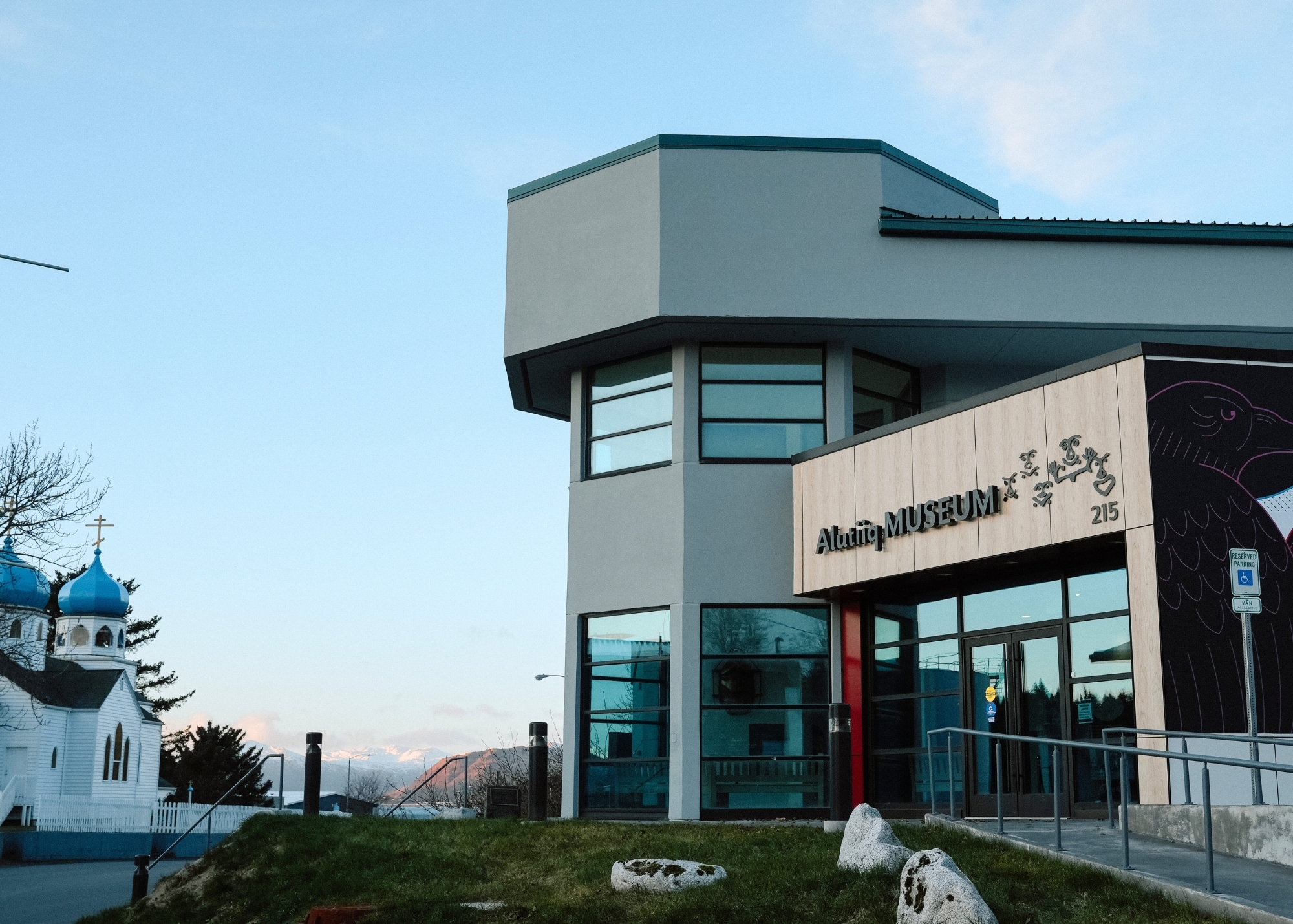 A modern gray building labeled Alutiiq Museum with large windows stands next to a white church with blue domes; mountains are visible in the background under a clear sky.