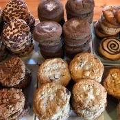 An assortment of cookies, including chocolate chip, double chocolate, peanut butter, pecan, oatmeal, and swirl cookies, are arranged in neat stacks on white trays on a wooden table.