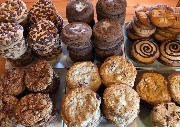 An assortment of cookies, including chocolate chip, double chocolate, peanut butter, pecan, oatmeal, and swirl cookies, are arranged in neat stacks on white trays on a wooden table.