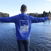 A person in a blue Kodiak, AK hoodie stands in shallow water with arms outstretched, facing a calm bay and rocky islands under a partly cloudy sky.