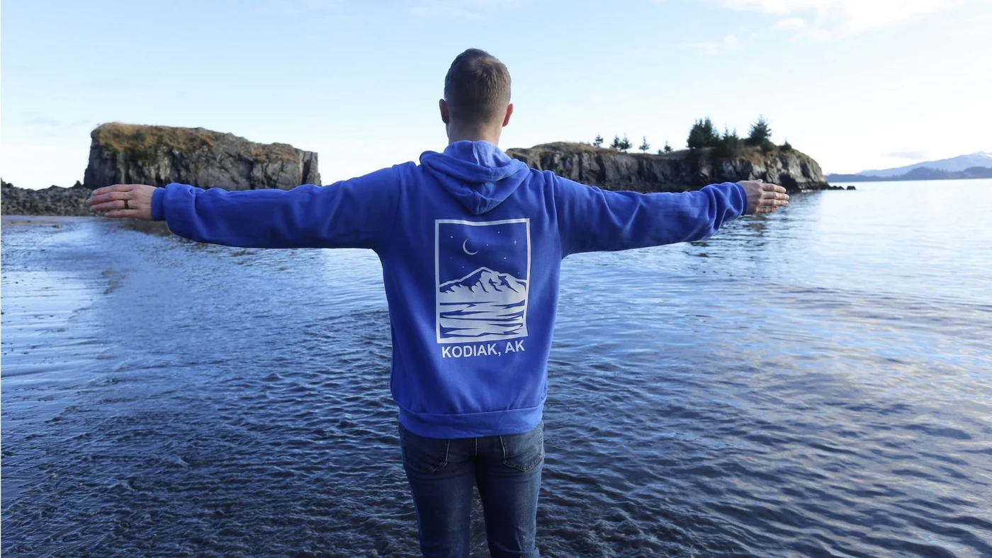 A person in a blue Kodiak, AK hoodie stands in shallow water with arms outstretched, facing a calm bay and rocky islands under a partly cloudy sky.