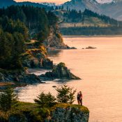 A couple stands on a cliff edge above the ocean at sunset, surrounded by pine trees, with mountains and forests in the background.