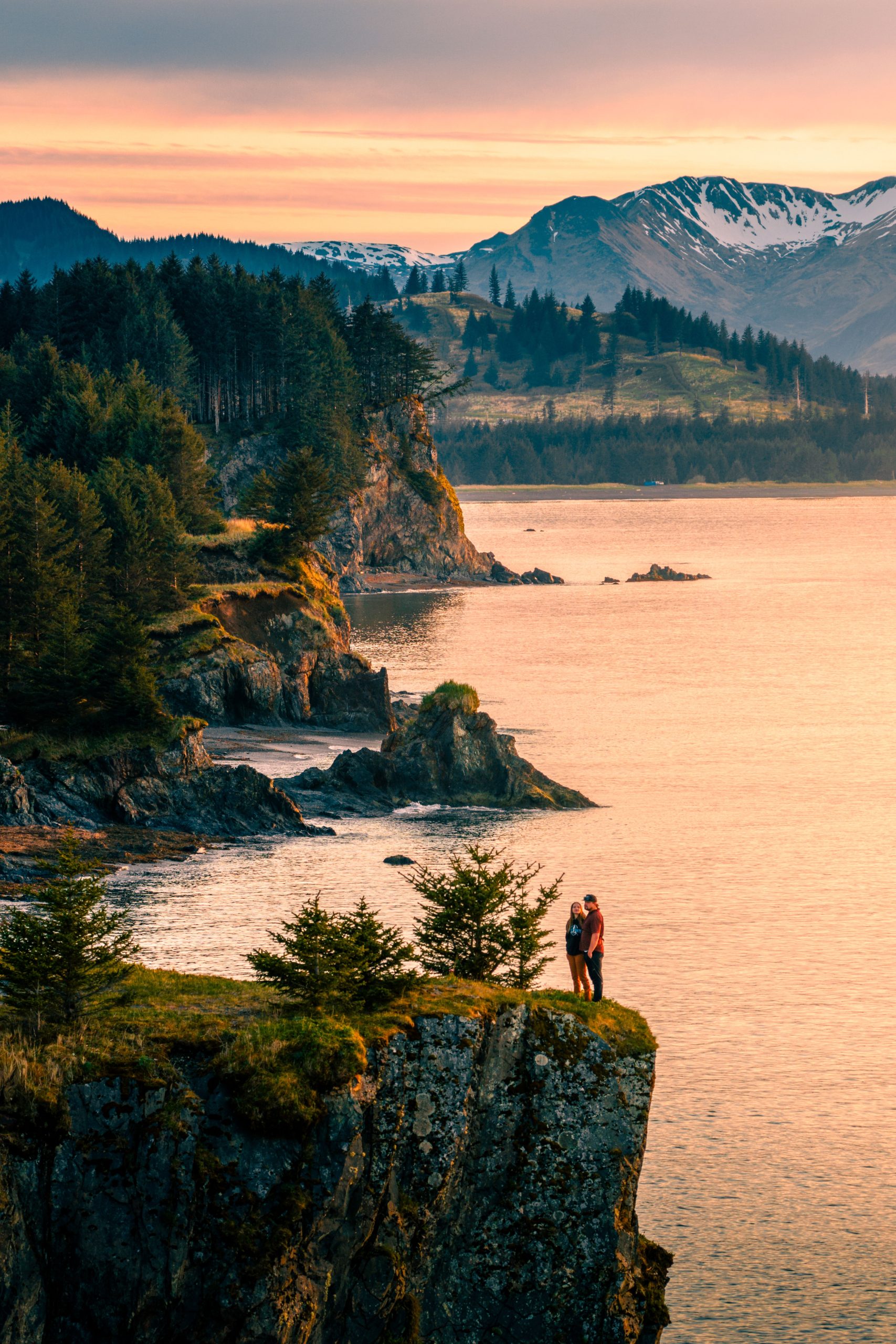 A couple stands on a cliff edge above the ocean at sunset, surrounded by pine trees, with mountains and forests in the background.
