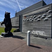 A large statue of a bear stands outside the Kodiak National Wildlife Refuge Visitor Center, which has a gray wooden exterior and a fish sculpture display on the wall. The sky is clear and sunny.