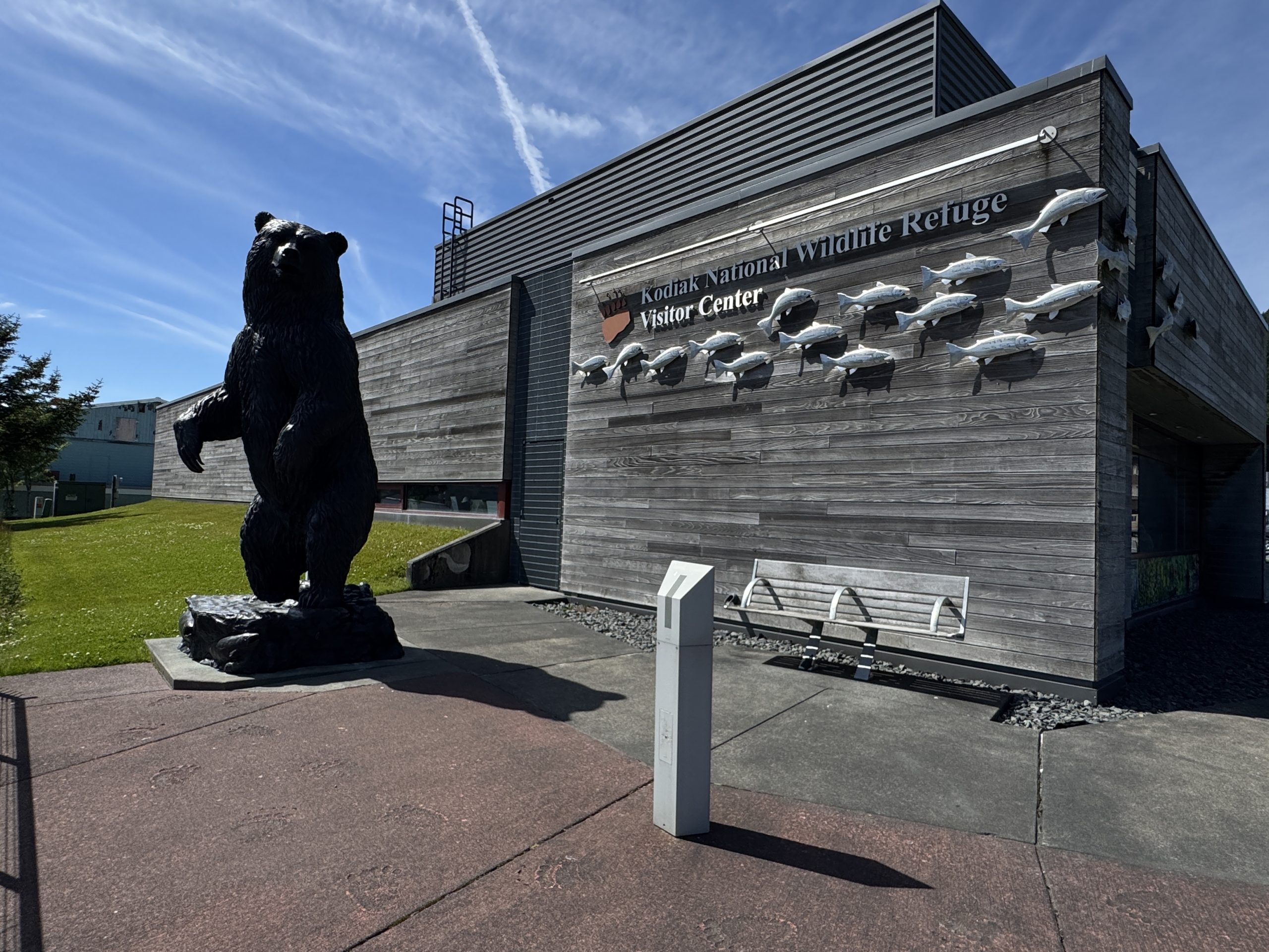 A large statue of a bear stands outside the Kodiak National Wildlife Refuge Visitor Center, which has a gray wooden exterior and a fish sculpture display on the wall. The sky is clear and sunny.