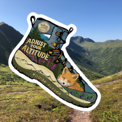 A mountainous landscape under a clear blue sky with a large illustrated hiking boot sticker overlay. The boot says, Adjust your altitude, Kodiak Alaska, and features flowers, mountains, and a fox inside.