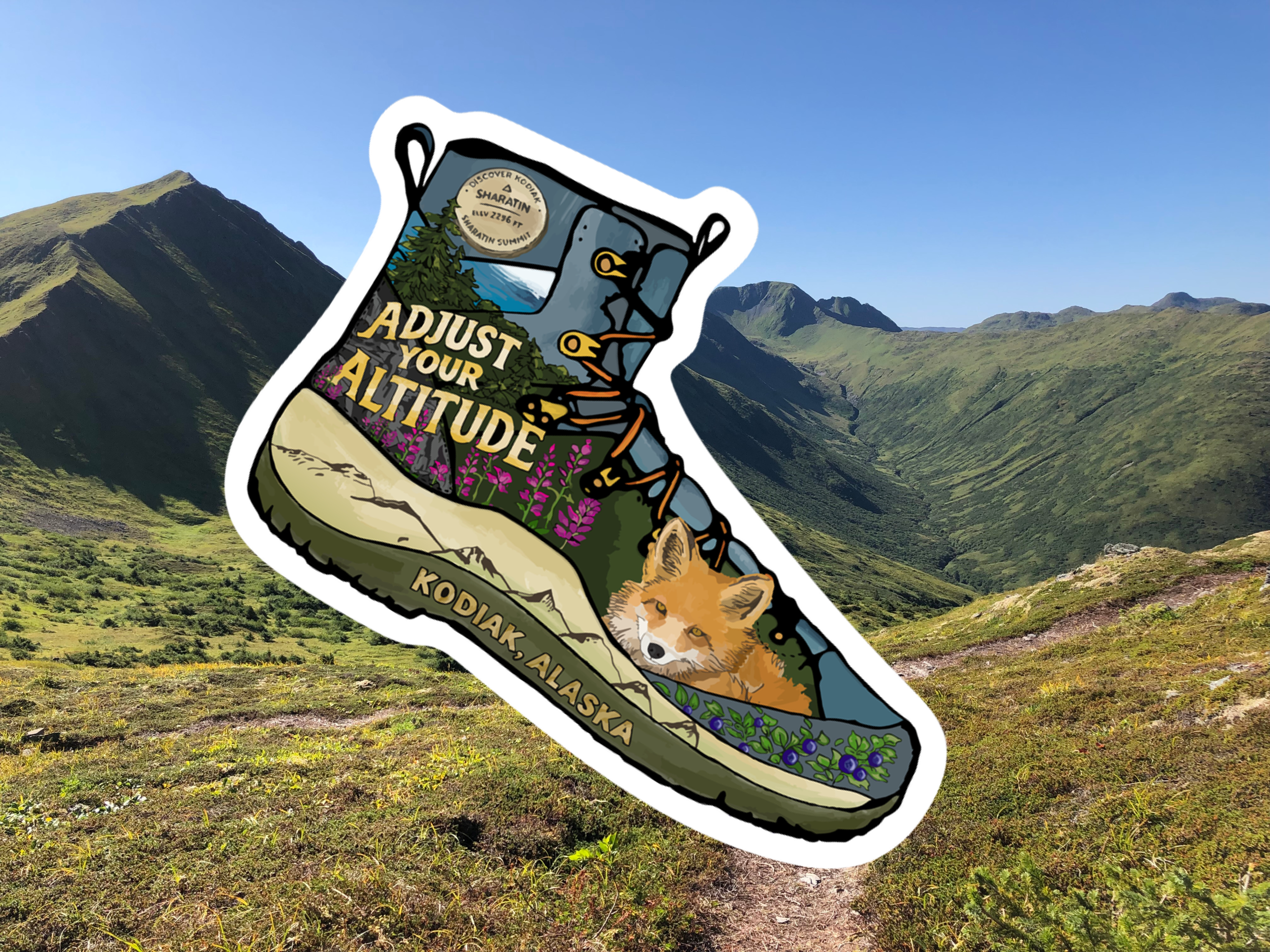A mountainous landscape under a clear blue sky with a large illustrated hiking boot sticker overlay. The boot says, Adjust your altitude, Kodiak Alaska, and features flowers, mountains, and a fox inside.