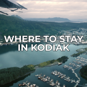 Aerial view of Kodiak, Alaska, showing a marina with boats, green hills, and a coastal town. The text Where to stay in Kodiak is placed in bold white letters over the center of the image.