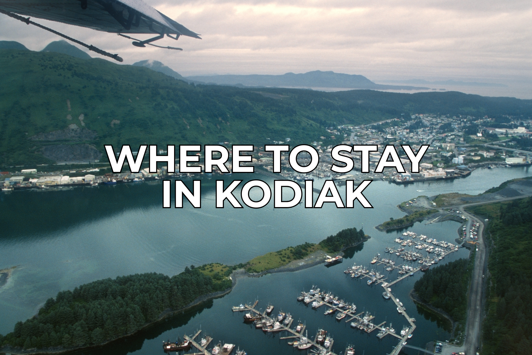 Aerial view of Kodiak, Alaska, showing a marina with boats, green hills, and a coastal town. The text Where to stay in Kodiak is placed in bold white letters over the center of the image.