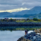 Two people stand on rocks by a calm body of water, fishing. In the background are rocky shores, grassy patches, a docked boat, and misty mountains under a cloudy sky.
