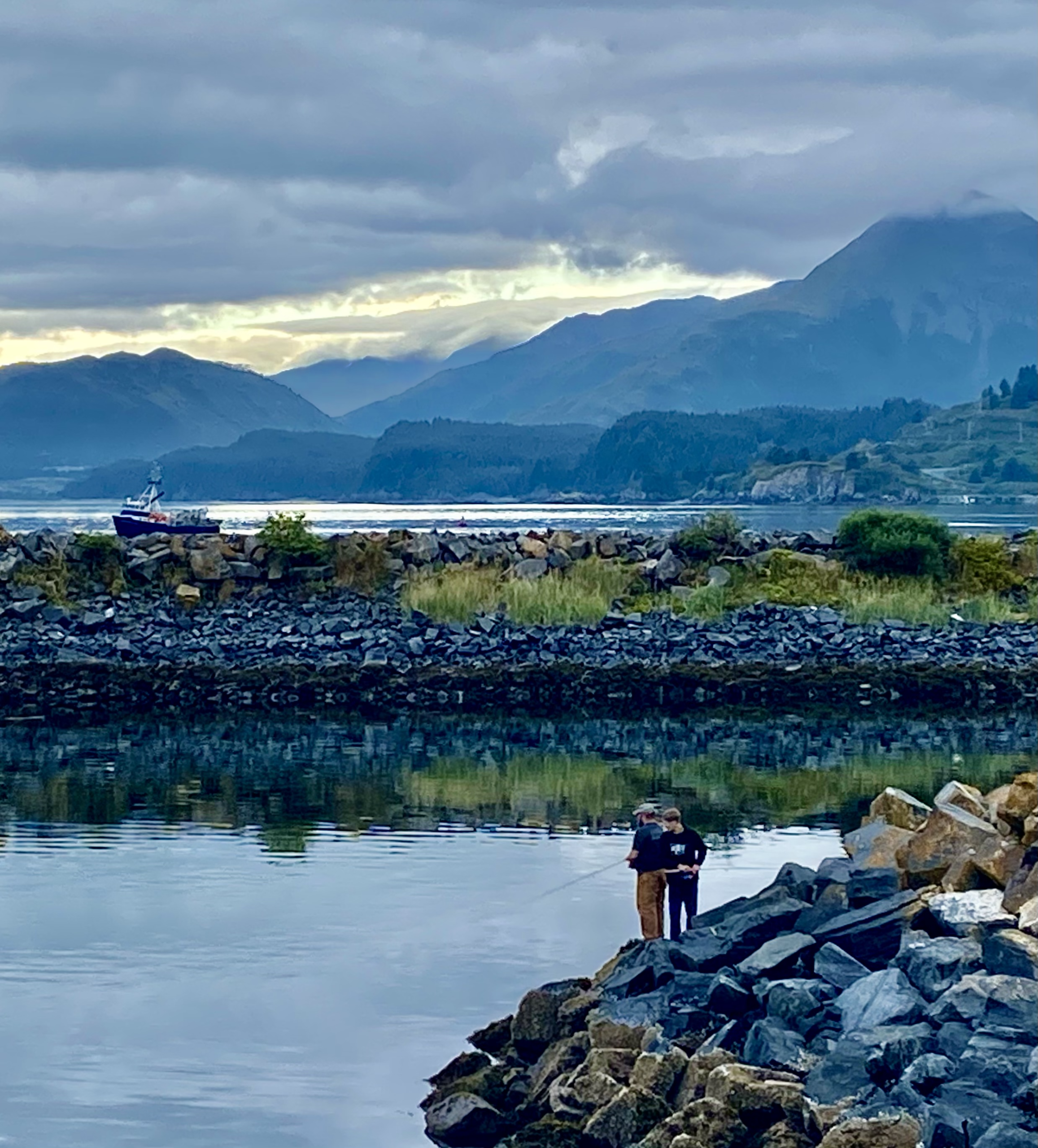 Two people stand on rocks by a calm body of water, fishing. In the background are rocky shores, grassy patches, a docked boat, and misty mountains under a cloudy sky.