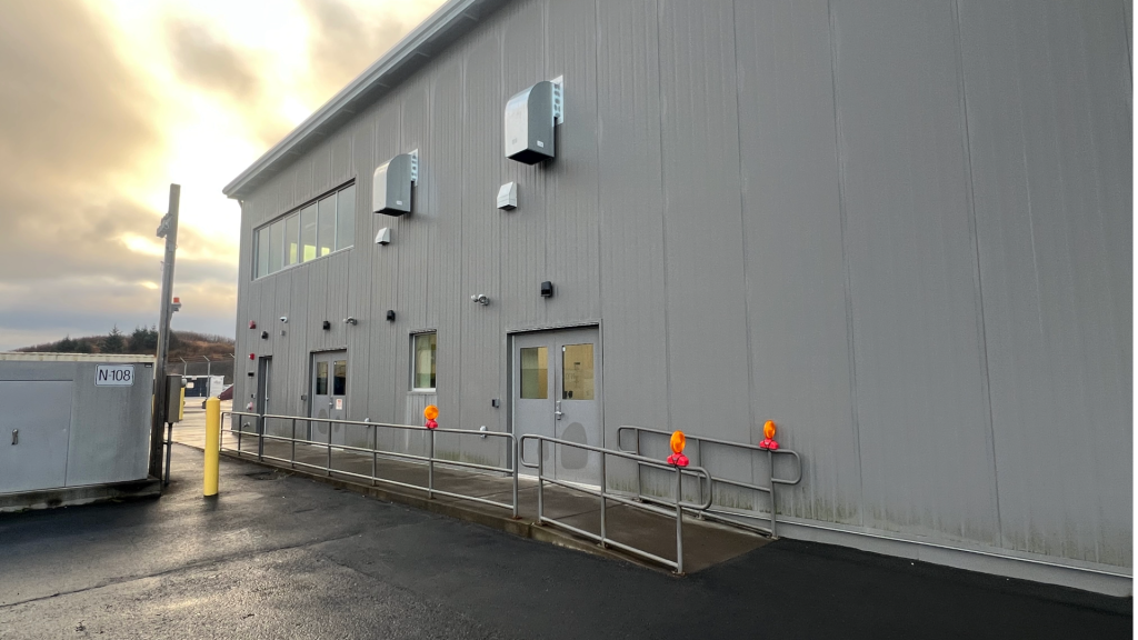 Exterior of a grey metal warehouse building serving as the Kodiak Airport temporary terminal, featuring two glass doors, a wheelchair ramp with metal railings, and a storage container marked N-108 in the foreground.