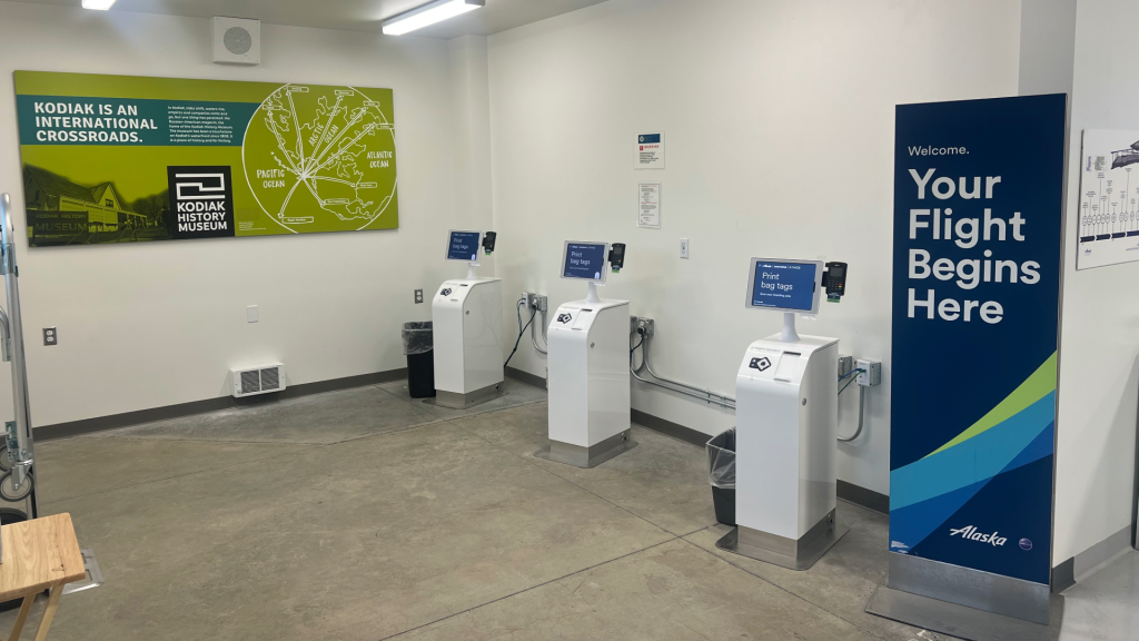 Three self-service bag tag kiosks stand in a row at the Kodiak Airport temporary terminal, positioned next to informational signs for the Kodiak History Museum and Alaska Airlines.